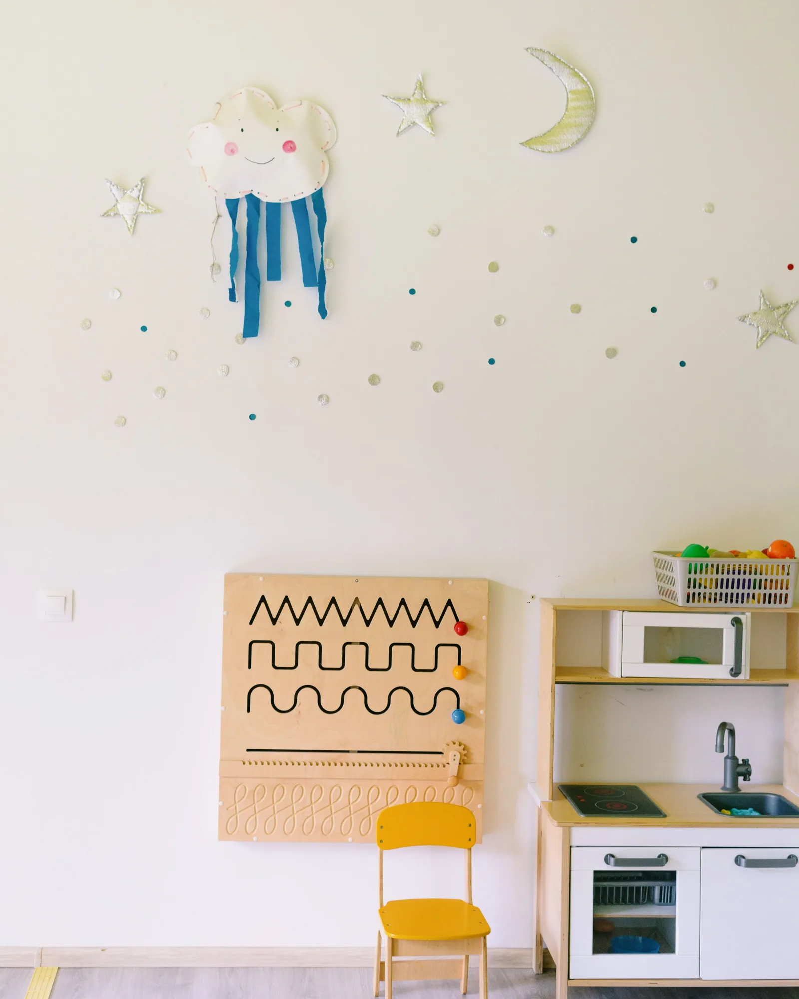 A tidy children's corner in a waiting area: a low wooden activity board, a small yellow chair and a toy kitchen against a pale wall with fabric cloud and star decorations.